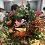 Ręcznie wykonany jesienny wianek ozdobiony zielonymi liśćmi, czerwonymi i żółtymi owocami, żołędziami oraz pomarańczowymi miechunkami. W tle widoczni są uczestnicy warsztatów.
